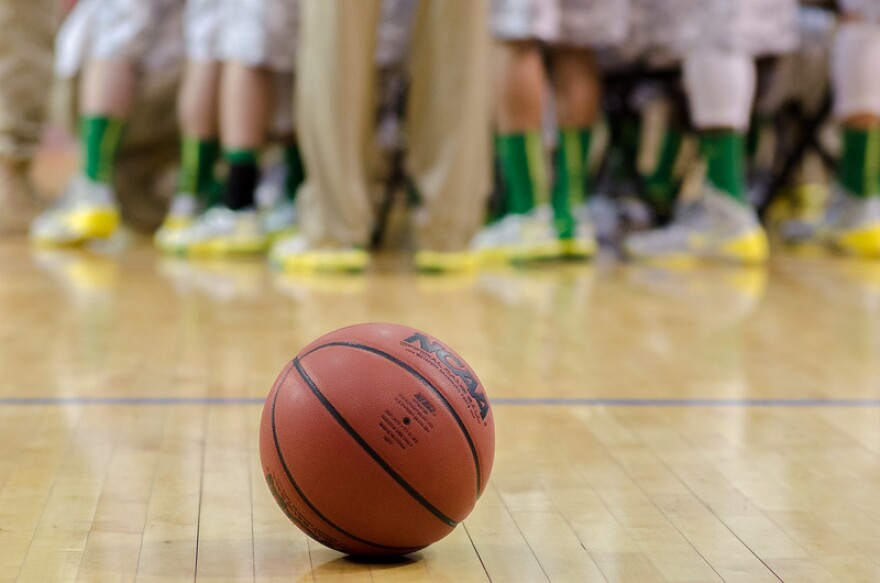 Basketball on a stadium floor with "NCAA" written on it and the feet of basketball players and a coach in the background.