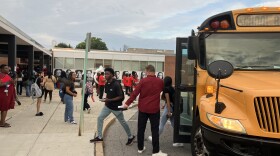 Woodlawn High School is pictured. Students are walking off of a yellow school bus. A marching band dressed in all red performs. 