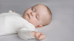 Calm silent adorable baby in white bodysuit sleeping on bed. Cute few month infant kid resting on pale sheet, linen. Child care, bedtime, daily routine concept. Close up