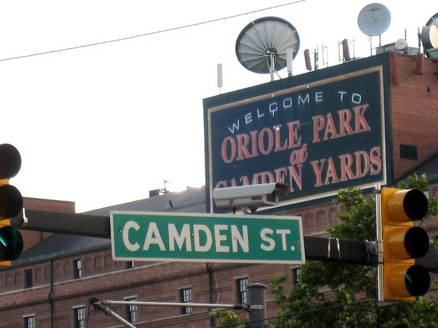  A street sign for Camden Street with a sign reading "Welcome to Oriole Park at Camden Yards" in the background on a brick building.