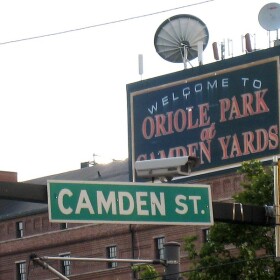  A street sign for Camden Street with a sign reading "Welcome to Oriole Park at Camden Yards" in the background on a brick building.