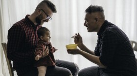 Male gay couple with adopted baby girl at home - Two handsome dads feed the baby girl on kitchen - Male babysitters - Lgbt family at home - Diversity concept